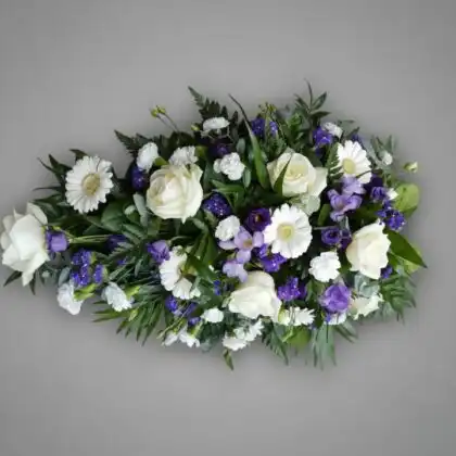 A casket topped with a serene arrangement of purple and white funeral flowers coffin sprays featuring lilies and roses.